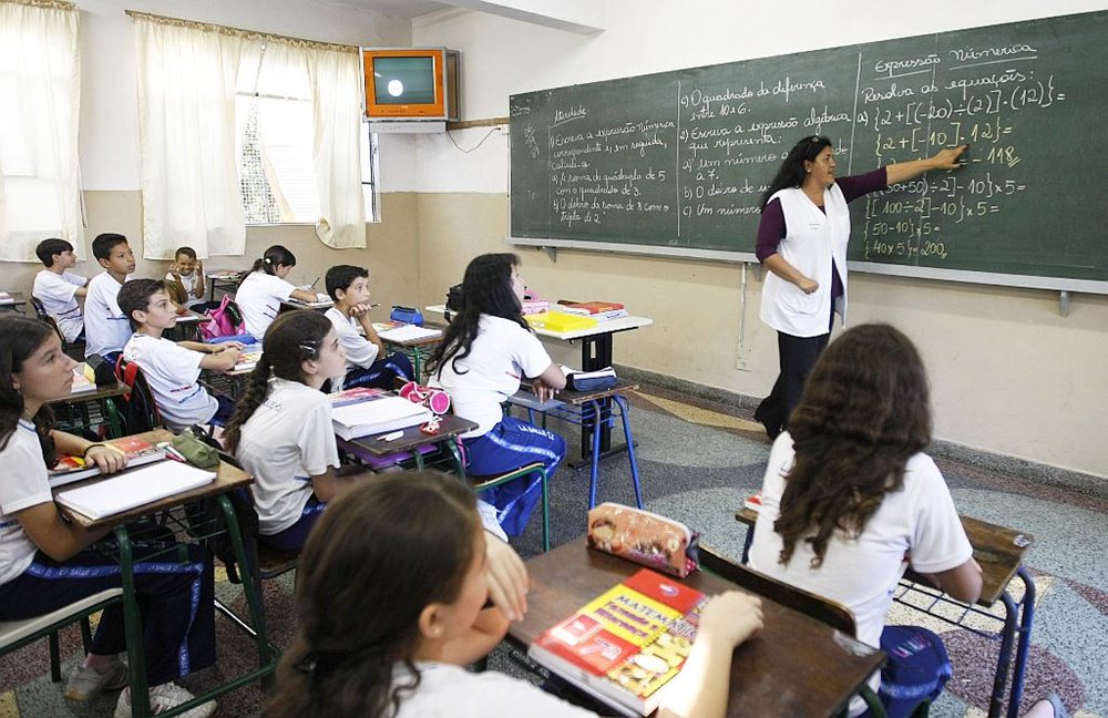 Alunos sentados em carteiras em sala de aula, olhando para o quadro negro, e professora dando aula