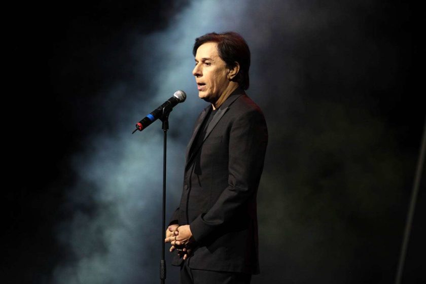 Tom Cavalcante no palco. Ele está falando em um microfone e usa terno completamente preto - Metrópoles