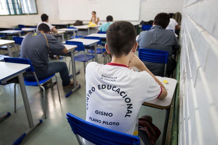 Aluno com uniforme sentado em carteira azul, de costas, e outros alunos em sala de aula ao fundo