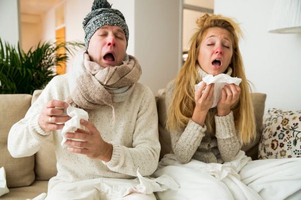 Fotografia colorida. Casal de homem e mulher aparece sentado em sofá com roupa de frio e espirrando