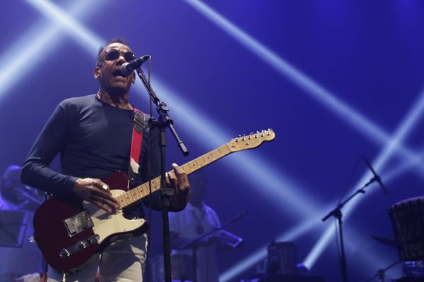 imagem colorida mostra cantor jorge ben jor em show. ele é negro, veste uma blusa azul escura e usa óculos escuros - metrópoles