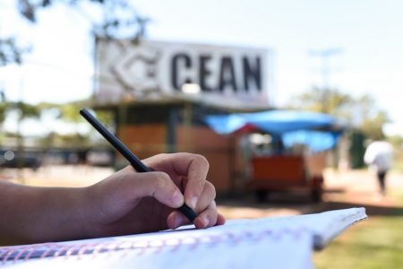 Mão segurando lápis e escrevendo em caderno e portão de escola ao fundo