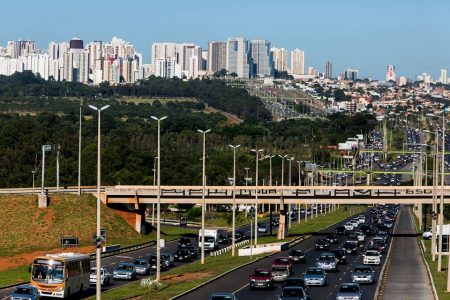 Pista com carros passando e viaduto e prédios ao fundo
