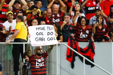 Homem segura cartaz branco com texto preto e torcida com uniforme preto e vermelho ao fundo