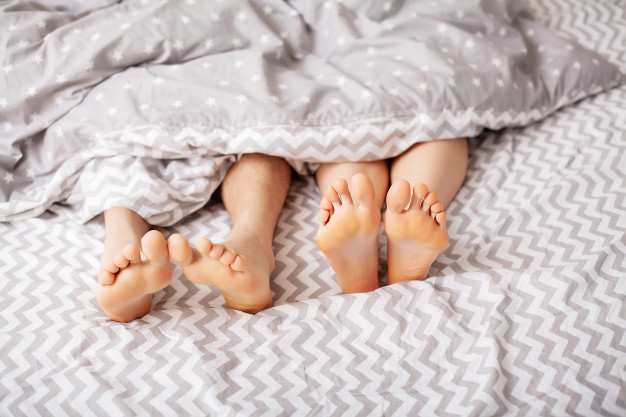 Dois pares de pés brancos em cima da cama saindo debaixo de um edredom cinza estampado