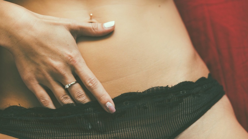 Foto colorida em close de uma mulher deitada sob um lençol vermelho colocando a mão por dentro da calcinha preta - Metrópoles