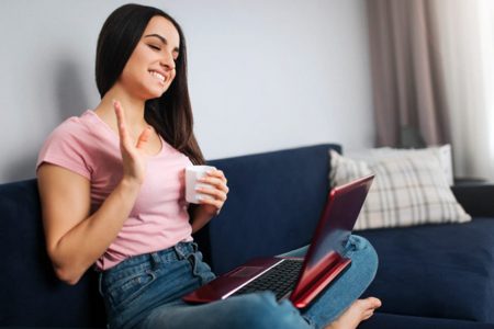 jovem-alegre-sentar-no-sofa-e-acenar-com-a-mao-ela-sorri-e-olha-na-tela-do-laptop-modelo-segura-lo-na-soneca-ela-tem-um-copo-branco-de-bebida_152404-4108