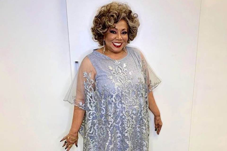 Foto colorida da cantora Alcione com um vestido lilás em um fundo branco - Metrópoles