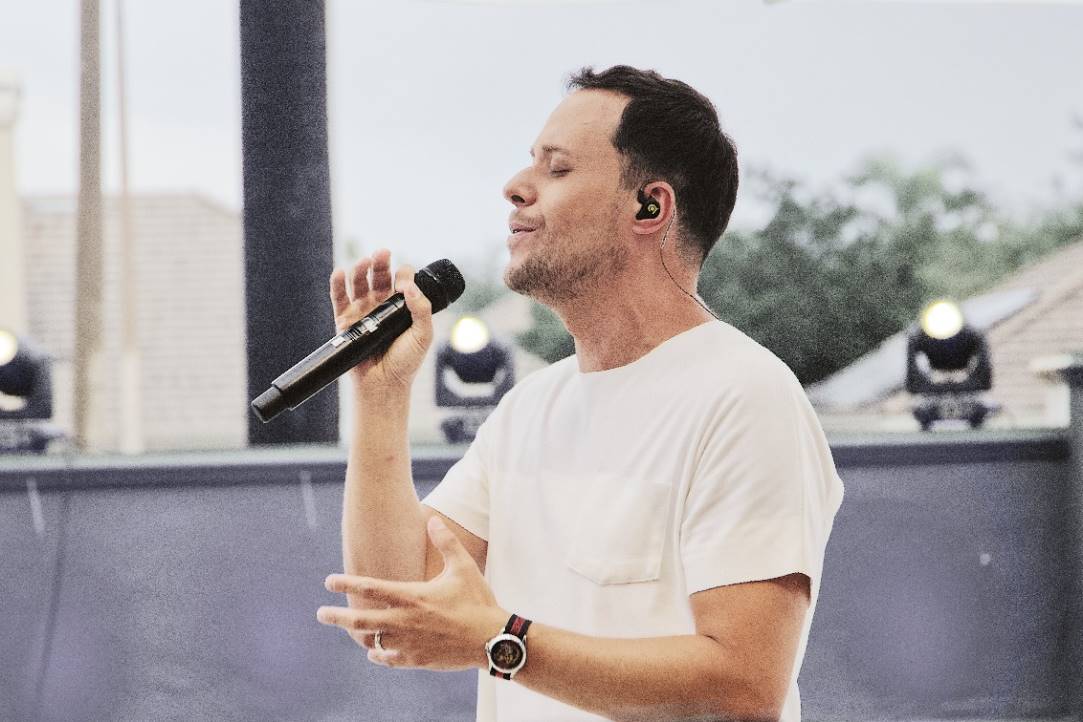 André Valadão está de roupa branca e segura um microfone em sua mão. Ele é branco, tem cabelo preto e está usando camisa branca e calça jeans