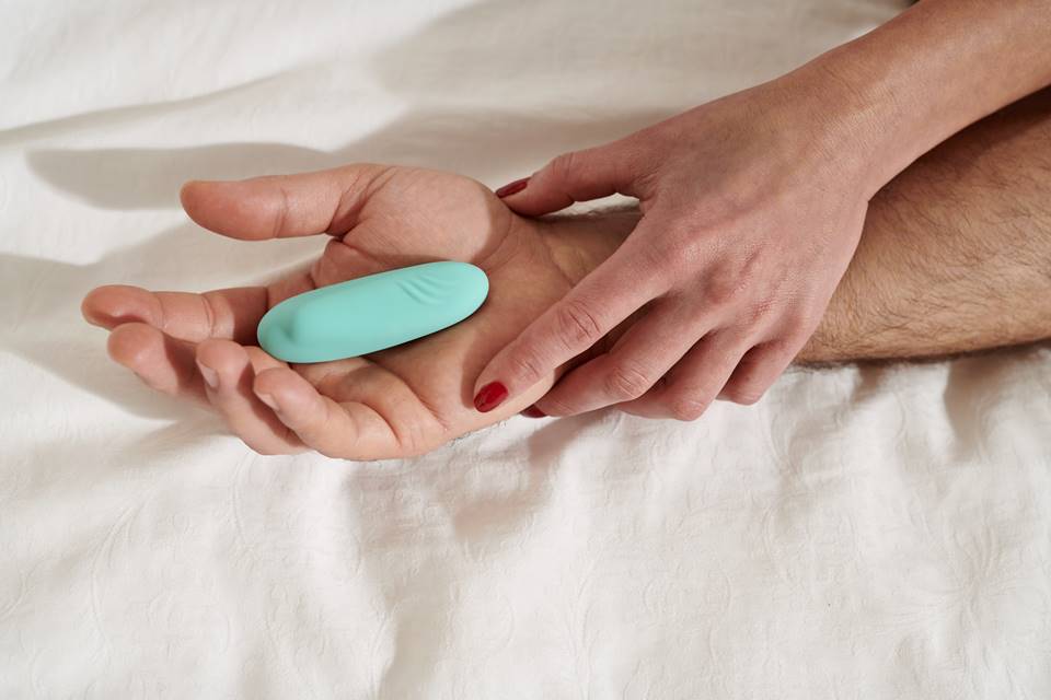 Foto colorida de mãos femininas segurando um vibrador azul claro em um fundo de lençol branco - Metrópoles