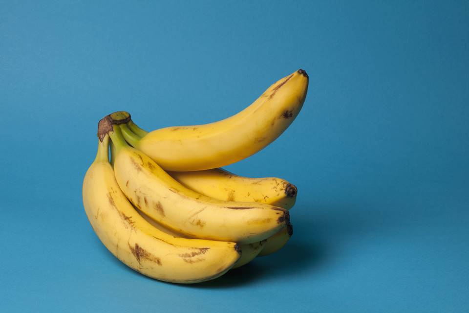 Foto colorida de um cacho de banana com uma das bananas levantadas em um fundo totalmente azul - Metrópoles