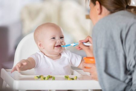 Na foto, um bebê branco sentado em uma cadeira sendo alimentado por uma mulher de costas - Metrópoles
