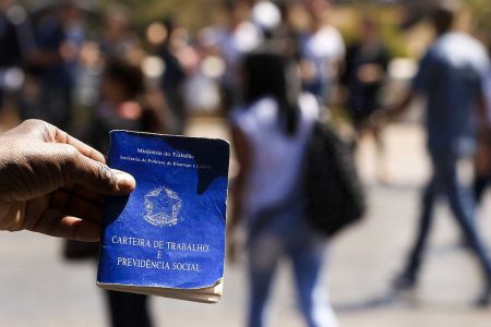 Mão segurando carteira de trabalho com pessoas caminhando na rua desfocadas ao fundo