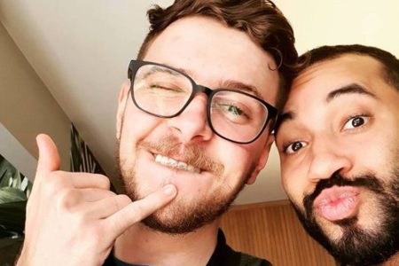 Gil do Vigor faz biquinho com a cabeça encostada em homem branco de óculos, que faz um hangloose com a mão
