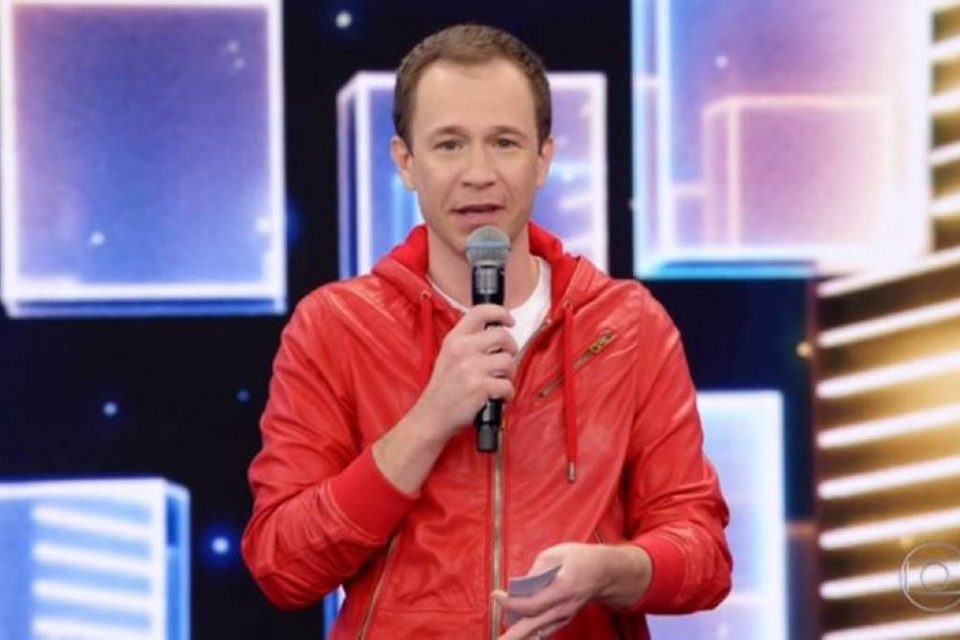 Tiago Leifert de blusa de frio laranja com microfone na mão no programa Domingão