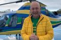 Fotografia colorida. Luciano posa para foto com jaqueta amarela. Ele está na frente de um helicóptero