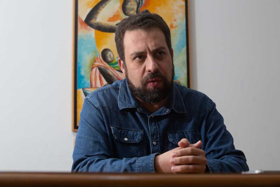 Imagem colorida mostra Guilherme Boulos, homem branco de cabelos e barba castanhos, de camisa jeans em uma sala