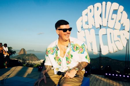 Foto colorida de Frrrugem com camisa estampada, sentado, com microfone