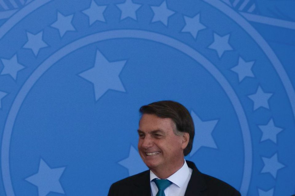 Presidente Jair Bolsonaro rindo; Ao fundo, escudo redondo azul-celeste, símbolo da república