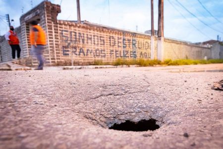 Imagem de enorme buraco em rua de Maceió. Ao fundo, um muro pichado com a seguinte frase: "Sonhos destruídos, éramos felizes" - Metrópoles