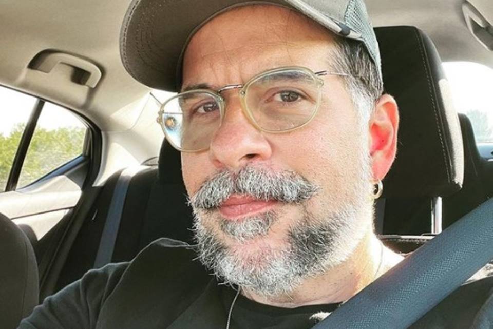 Foto colorida de Leandro Hassum. O ator está de boné, barba grisalha e blusa preta. Ele está em um carro e usa cinto de segurança - Metrópoles