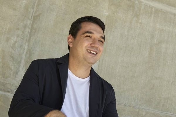 Na foto, vemos um homem de camiseta branca e blazer preto olhando para longe e sorrindo
