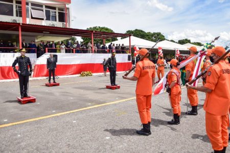 Homens fardados com roupa laranjada. Eles seguram armas e estão de frente para homens com roupas pretas