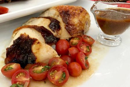 Na foto, um frango assado fatiado com tomates cereja e molho marrom em cima de um prato branco