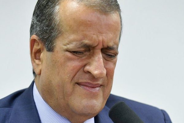 Fotografia colorida. Valdemar aparece no centro da imagem, vestindo paletó azul com gravata da mesma cor. Ele aparenta fazer uma careta