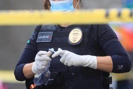 Fotografia colorida. Legista aparece no fundo da imagem em uma cena de crime