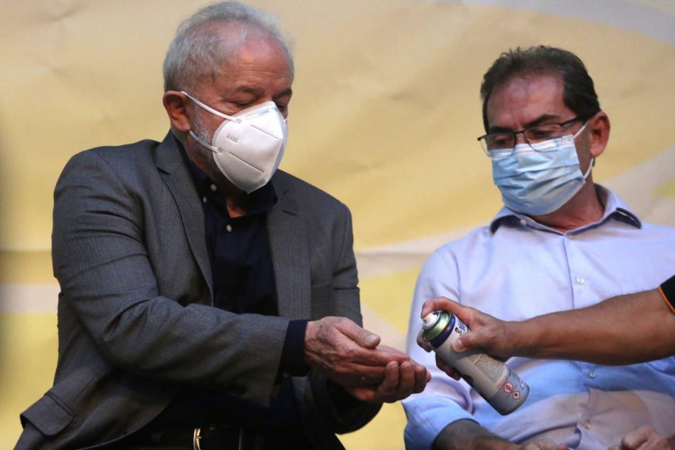 Ex-presidente Lula em foto ao lado de Paulinho da Força (Solidariedade). Ambos usam máscara, roupa social e uma mão oferece álcool em spray para Lula, enquanto Paulinho olha - Metrópoles