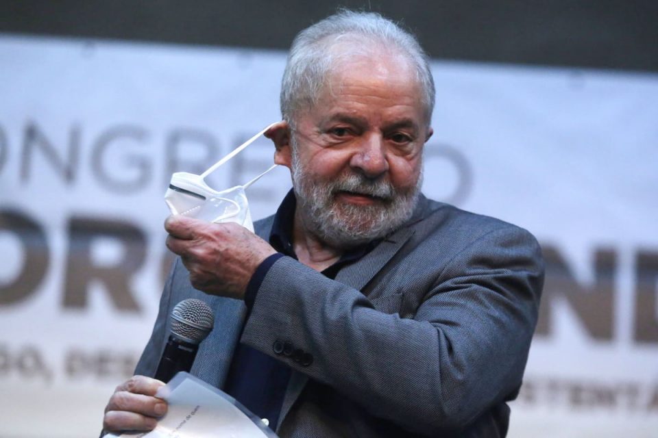 Lula tira a máscara para falar em evento. Ele segura um microfone e usa terno - Metrópoles