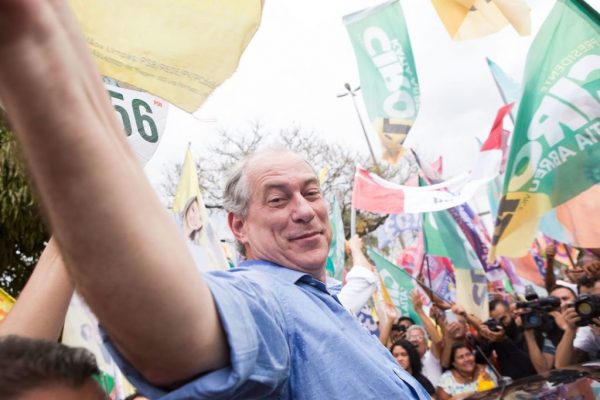 Fotografia colorida de Ciro Gomes. Ele é um homem branco, de cabelos lisos e grisalhos penteados para o lado. Na foto, aparece de braços abertos, em meio a uma multidão.