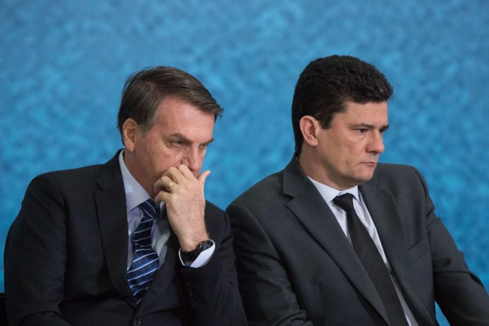 Na imagem colorida, dois homens estão centralizados. Eles usam terno e gravada escuro, possuem cabelo curto e ambos são brancos