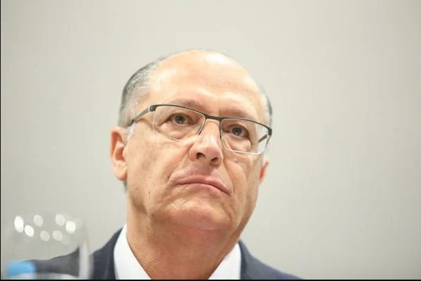 Na imagem colorida, um homem está posicionado no centro da imagem. Ele usa blazer preto e camiseta branca