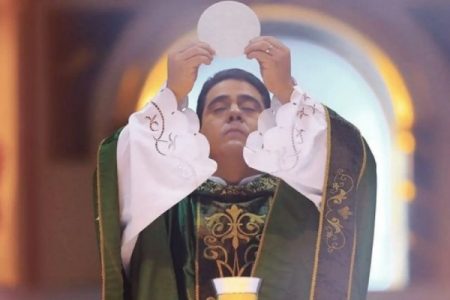 Na fotografia colorida, padre Robson está celebrando missa e levanta a comunhão para o alto de olhos ffechados