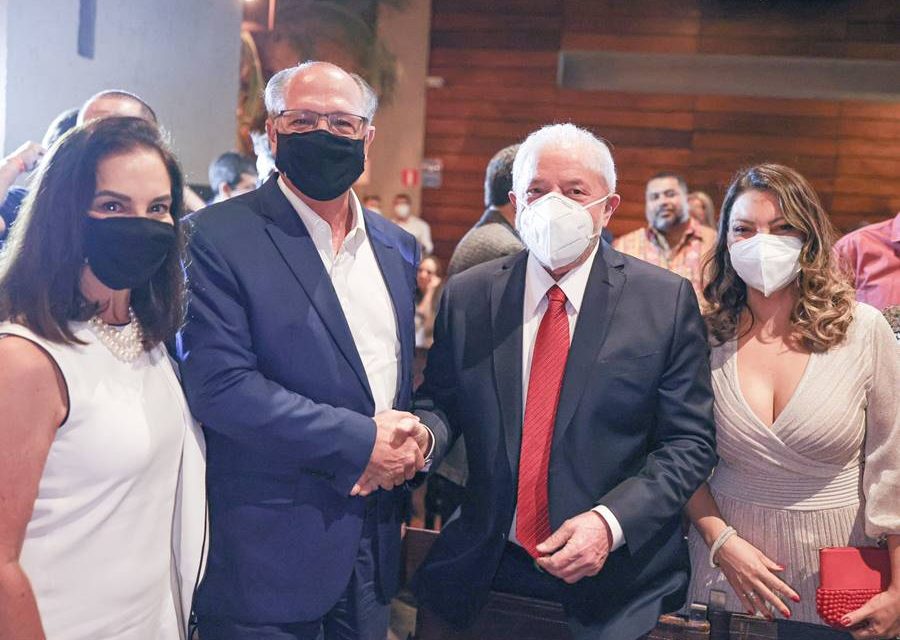 Foto colorida de encontro do ex-presidente Lula com Geraldo Alckmin. Na imagem, os dois aparecem olhando para a câmera, apertando as mãos.