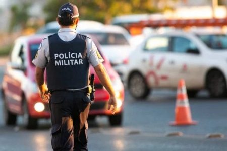 Homem com farda da Polícia Militar de costas e carros no trânsito ao fundo