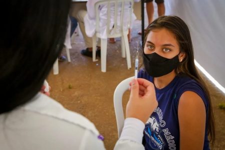 Na imagem colorida, uma criança está de frentre para uma enfermeira com uma seringa nas mãos