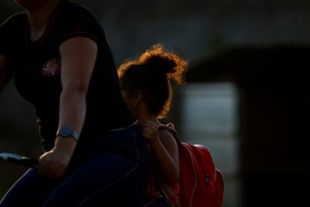 mulher e criança de costas, andando de bicicleta. A criança, com uma mochila vermelha nas costas segurando a blusa preta da mulher que direciona a bicicleta