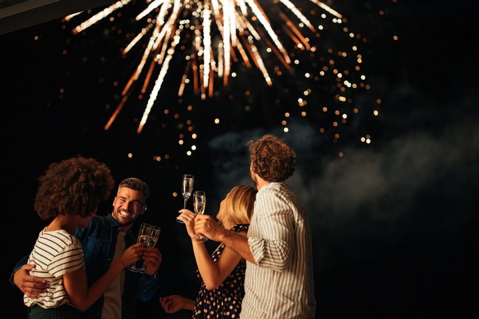 Grupo de quatro amigos. Eles estão com taça na mão e olham a queima de fogos de artifício