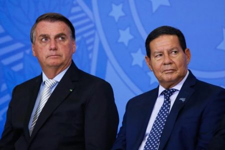 Na imagem colorida, dois homens estão posicionados no centro. Eles usam terno e gravata na cor preta e camiseta branca. Eles tem cabelos curtos e escuros