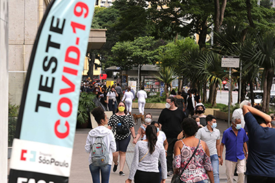 Pessoas caminhando na rua e placa ao lado escrito "Teste Covid-19"