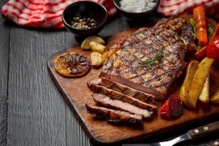 Na foto, carne grelhada rodeada de legumes em cima de uma tábua de madeira