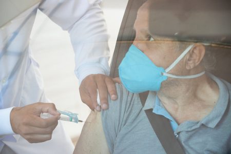 Pessoa de máscara dentro do carro e mão aplicando vacina no braço