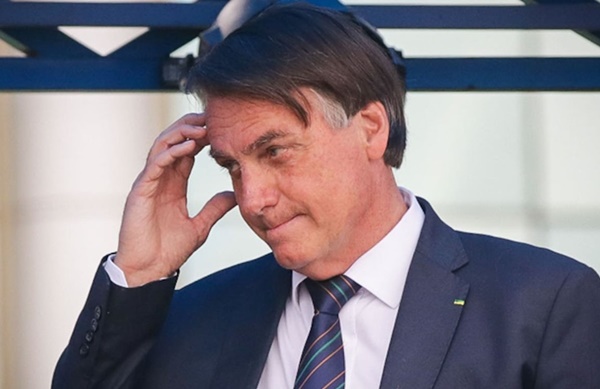 Na imagem colorida, um homem está posicionado no centro. Ele uda terno e gravata em cor azul escuro, camiseta branca e está com a mão na cabeça enquanto olha para o lado inferior esquerdo