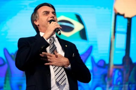 Na imagem colorida. Um homem está posicionado no centro. Ele usa terno e gravata, tem cabelos curtos e grisalhos e segura microfone com as mãos. Ele está discursando