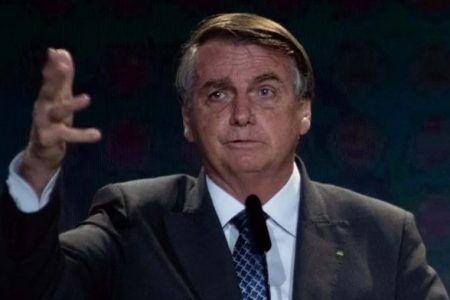 Na imagem colorida. Um homem está posicionado no centro. Ele usa terno e gravata, tem cabelos curtos e grisalhos e olha atentamente para a frente