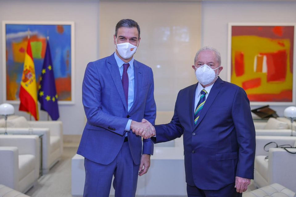 Lula cumprimenta o primeiro-ministro da Espanha, Pedro Sánchez, durante viagem à Europa. Eles estão em salão branco amplo, com quadros e a bandeira da Espanha ao lado da bandeira da União Européia, ambos e terno, e apertando as mãos - Metrópoles