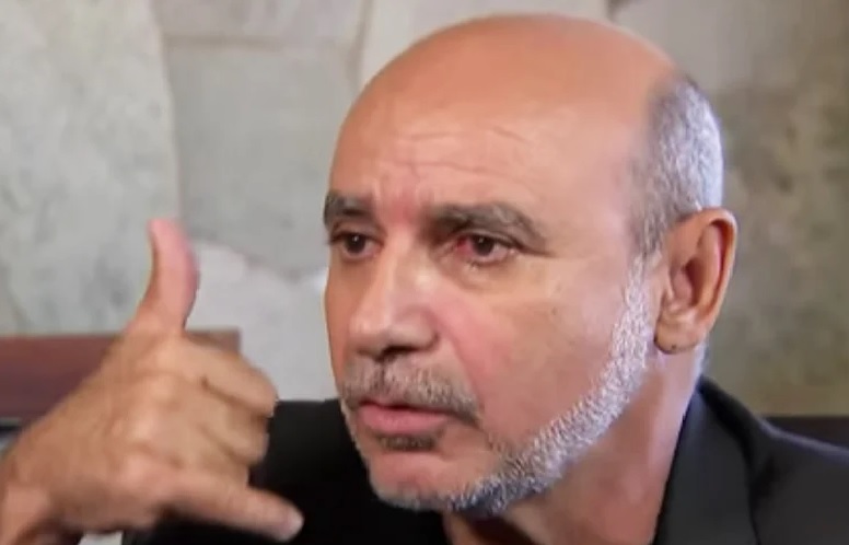 Na imagem colorida, um homem está posicionado no centro. Ele é careca e está com as mãos levantadas